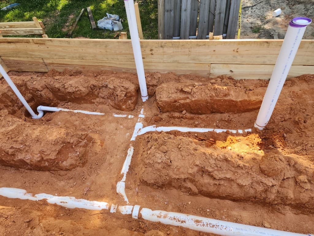 White PVC plumbing rough-in in dug trenches with multiple pipe runs and vertical risers at a residential site